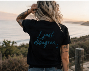 A woman with blonde hair stands with her back to the camera wearing a black t-shirt with the text 'I just disagree.' printed in teal on the back, set against a coastal sunset background.