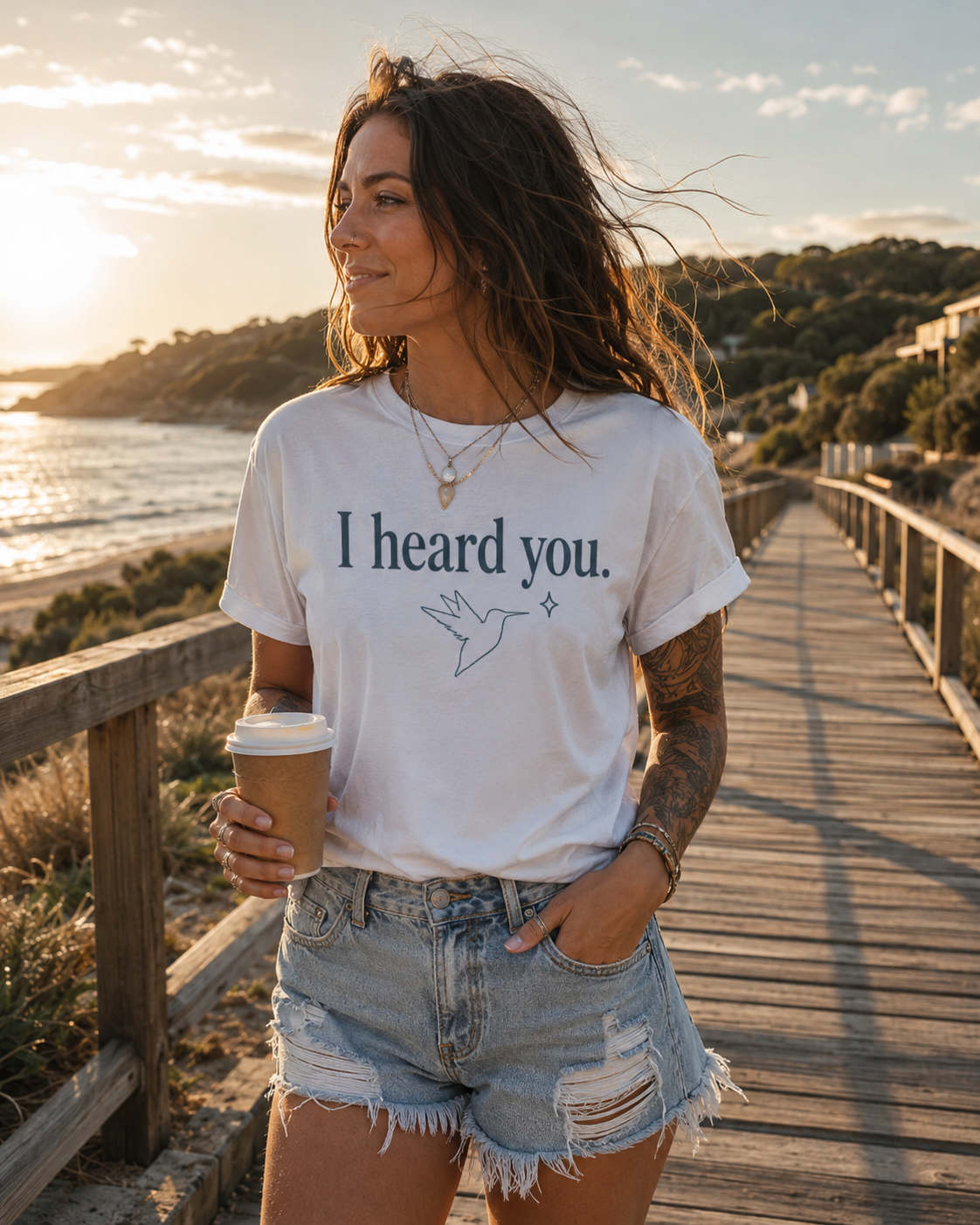A woman stands on a wooden boardwalk by the ocean at sunset wearing a white graphic T-shirt that reads 'I heard you.' with a dove illustration, paired with distressed denim shorts and holding a takeaway coffee cup.