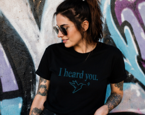 A woman with tattoos and sunglasses wearing a black t-shirt with a teal graphic that reads 'I heard you.' and a shark silhouette, posed against a graffiti-covered wall.