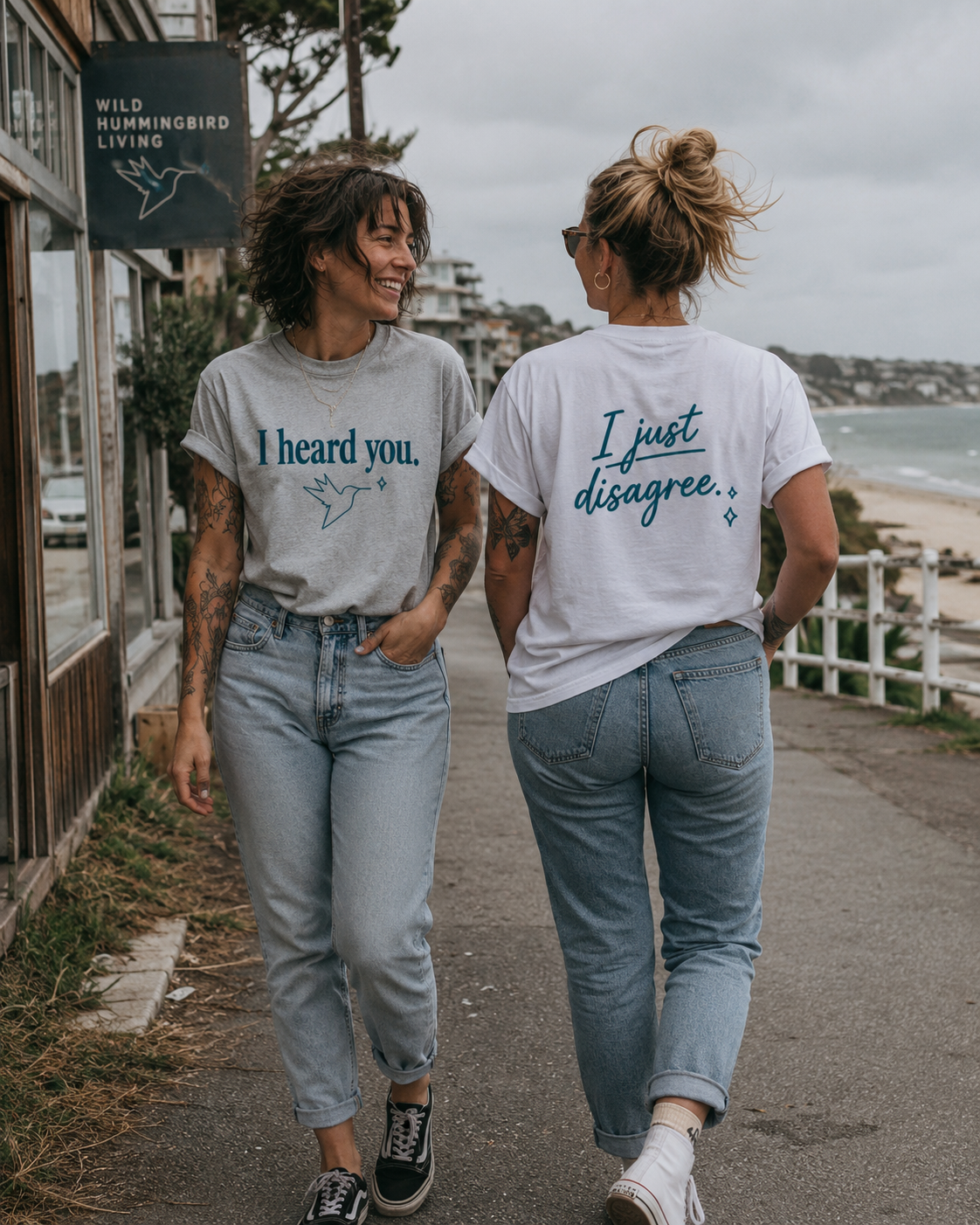 Two women walking away on a seaside path, wearing matching graphic t-shirts. The woman on the left wears a light gray t-shirt with 'I heard you.