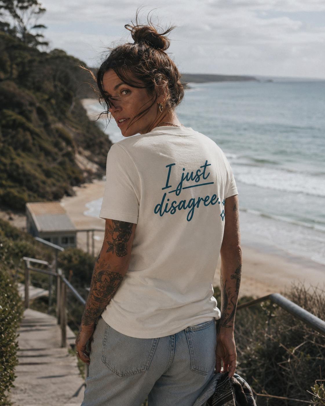 A woman with tattoos stands on a coastal pathway wearing a cream-colored t-shirt with the text 'I just disagree.' printed in dark blue on the back, paired with light blue jeans, with a beach and ocean in the background.
