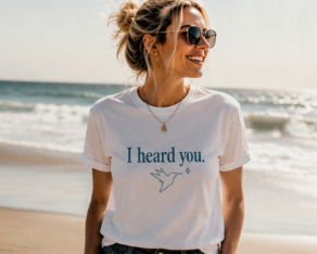 A woman wearing a white graphic t-shirt with the text 'I heard you.' and a bird illustration, standing on a beach with the ocean in the background.
