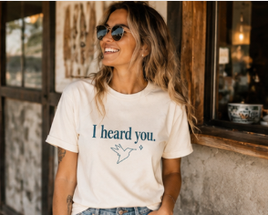 A woman wearing a cream-colored graphic t-shirt with the text 'I heard you.' and a small bird silhouette printed in dark blue.